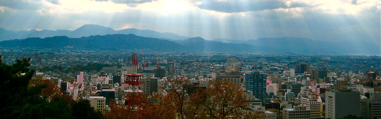 The sun peaks through the clouds to show a city with many tall buildings in front of a series of mountains. A tall red and white tower and a Ferris Wheel are also visible. 